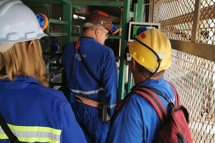 Tres personas con equipo de minería de pie junto a una puerta o estructura metálica verde.