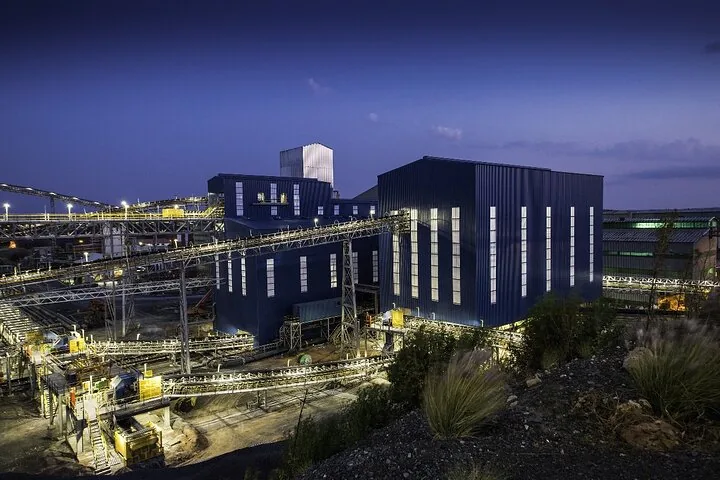 Struttura mineraria industriale illuminata e nastri trasportatori contro un cielo crepuscolare.